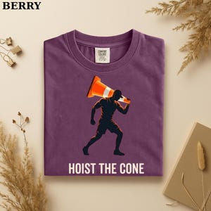 May include: Berry-colored t-shirt with a graphic of a person carrying an orange and white traffic cone, with the text "HOIST THE CONE" in white. The shirt has a crew neck and short sleeves. The brand name "BERRY" is at the top left.