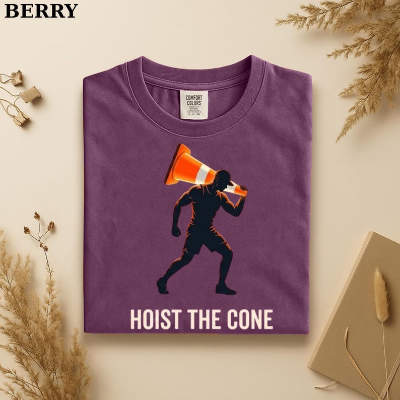 May include: Berry-colored t-shirt with a graphic of a person carrying an orange and white traffic cone, with the text "HOIST THE CONE" in white. The shirt has a crew neck and short sleeves. The brand name "BERRY" is at the top left.