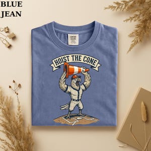 May include: A blue t-shirt with the text "HOIST THE CONE" above a cartoon baseball player eagle holding a traffic cone. The eagle is wearing a baseball uniform and cap. The shirt is made of a soft material.