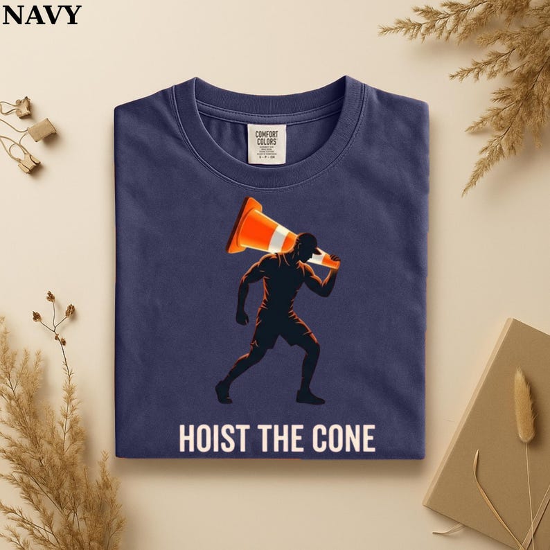 May include: Navy blue t-shirt with a graphic of a person carrying an orange traffic cone. The text "HOIST THE CONE" is printed in white below the graphic. The shirt has a crew neck and short sleeves.