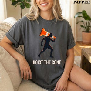 May include: Dark gray t-shirt featuring a graphic of a person carrying a traffic cone. The person is in silhouette, with the cone in orange and white. The text "HOIST THE CONE" is printed below the graphic in white.