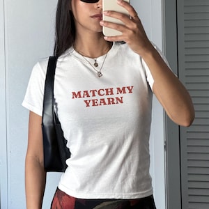 Op de afbeelding: Wit T-shirt met de tekst "MATCH MY YEARN" in het rood. Het shirt heeft korte mouwen en een ronde hals. Een zwarte schoudertas is zichtbaar aan de linkerkant.