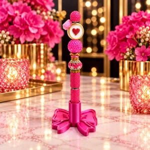 May include: A vertical pen stand featuring a bright pink bow-shaped base. The pen is decorated with stacked beads in shades of pink, including a round white charm with a red heart, a textured pink sphere, and a gold-toned bead with pink accents.