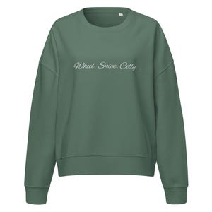 May include: A sage green sweatshirt with the text "Wheel. Snipe. Celly." in a cursive font. The sweatshirt has a crew neck and long sleeves with ribbed cuffs and hem. The fabric appears to be a soft, comfortable material.