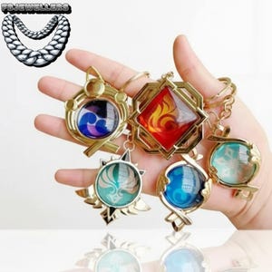 May include: A selection of gold-coloured keychains with diverse designs. Each keychain features a gemstone or glass insert in different colours, including blue, red, and green. The keychains have detailed metalwork and are held in a hand.