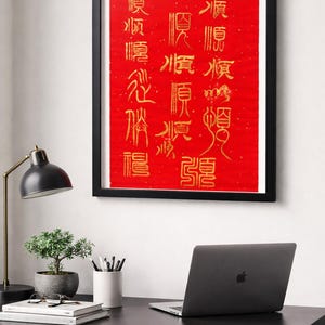 Puede incluir: Una obra de arte roja enmarcada con caligrafía china dorada. El marco negro rodea el fondo rojo con texto vertical. La obra de arte se exhibe en un escritorio negro con una computadora portátil, una pequeña planta en maceta y una lámpara de escritorio.