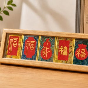 Puede incluir: Un marco de madera rectangular muestra cinco paneles con caracteres chinos rojos. Los paneles tienen fondos coloridos y estampados. El marco está apoyado sobre una superficie de madera, con un jarrón y libros al fondo.