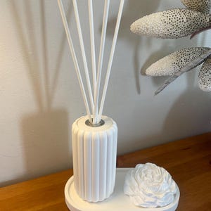 May include: A white reed diffuser set featuring a cylindrical, vertically ribbed bottle with five white diffuser sticks inserted into the top. The bottle sits on a white oval tray alongside a white, sculpted flower-shaped decorative object.