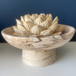 May include: A pedestal bowl featuring a cream and tan marbled pattern with dark brown veining. Resting inside the bowl is a sculpted, multi-layered flower ornament in a solid cream colour, characterised by pointed, overlapping petals arranged in a circular formation.
