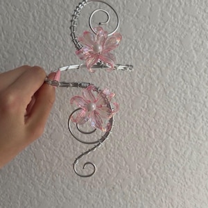 May include: A silver-toned wire ear cuff featuring two pink translucent flower-shaped beads with white centers. The wire is coiled into decorative swirls that extend along the length of the cuff, creating a structured and ornate design.