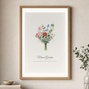 Lifestyle room mockup showing a personalized birth flower bouquet in a light wood frame above a warm neutral console. The watercolor family flower art includes individual birth month flowers, names and the custom title Mom's Garden. Soft daylight, a ceramic vase and simple decor show how this Christmas gift for mom or grandma looks in a calm modern home.
