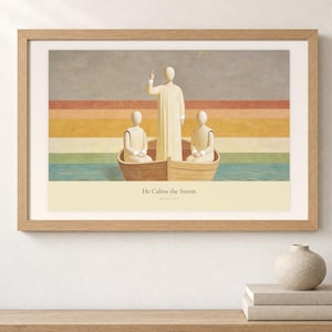 Shelf mockup of He Calms the Storm in a light oak frame resting on a pale wood surface beside stacked neutral books and a rounded ceramic vase. The framed print shows Christ standing in a wooden boat with a raised hand above calm layered water. Cream matting, soft wall texture, and natural light present the horizontal Bible artwork at an easy domestic scale.