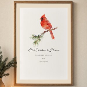 Lifestyle holiday room mockup showing a personalized First Christmas in Heaven print in a warm wood frame above a softly decorated console. A red cardinal rests among snow covered pine branches above Margaret Johnson and 2026. Gentle lights, evergreen accents and neutral decor show how this memorial Christmas gift brings a quiet remembrance into a family home.