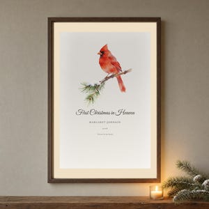Hero framed view of a personalized First Christmas in Heaven memorial print. The artwork features a bright red cardinal perched among snow covered pine needles, with Margaret Johnson and the year 2026 set clearly below the seasonal message. Clean glass, a natural wood frame and soft winter light reveal the delicate botanical details and calm paper texture of the tribute.