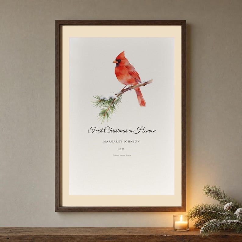 Hero framed view of a personalized First Christmas in Heaven memorial print. The artwork features a bright red cardinal perched among snow covered pine needles, with Margaret Johnson and the year 2026 set clearly below the seasonal message. Clean glass, a natural wood frame and soft winter light reveal the delicate botanical details and calm paper texture of the tribute.
