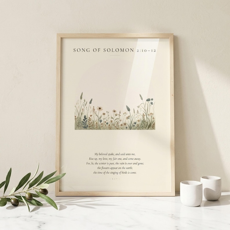 Custom Wildflowers watercolor scripture print in thin natural birch frame with clean glass front showing diagonal reflection, leaning on white Carrara marble surface next to a fresh olive branch with green leaves and two small white ceramic votive holders, bright morning light, cottage Boho still-life styling for Christian wedding wall art digital download from TheFramedVerse