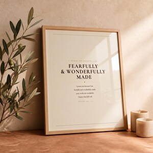 Botanical glass frame scene with the artwork near leafy stems, gentle reflections, warm wood, and soft natural light. The Fearfully and Wonderfully Made artwork uses minimalist boho typography in beige, brown, and black. This Psalm 139:14 print suits a nursery, baptism gift, baby dedication, bedroom, or quiet Christian reading corner as printable digital wall art.