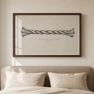 Cord of Three Strands wall art in a walnut frame hanging above a minimalist bed with cream linen pillows and oat headboard, soft window-light reflection on the glass. Christian Ecclesiastes 4:12 wedding scripture print, instant digital download for anniversary or marriage gifts.