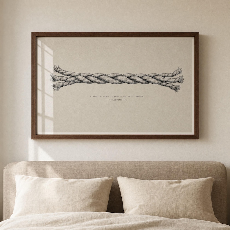 Cord of Three Strands wall art in a walnut frame hanging above a minimalist bed with cream linen pillows and oat headboard, soft window-light reflection on the glass. Christian Ecclesiastes 4:12 wedding scripture print, instant digital download for anniversary or marriage gifts.