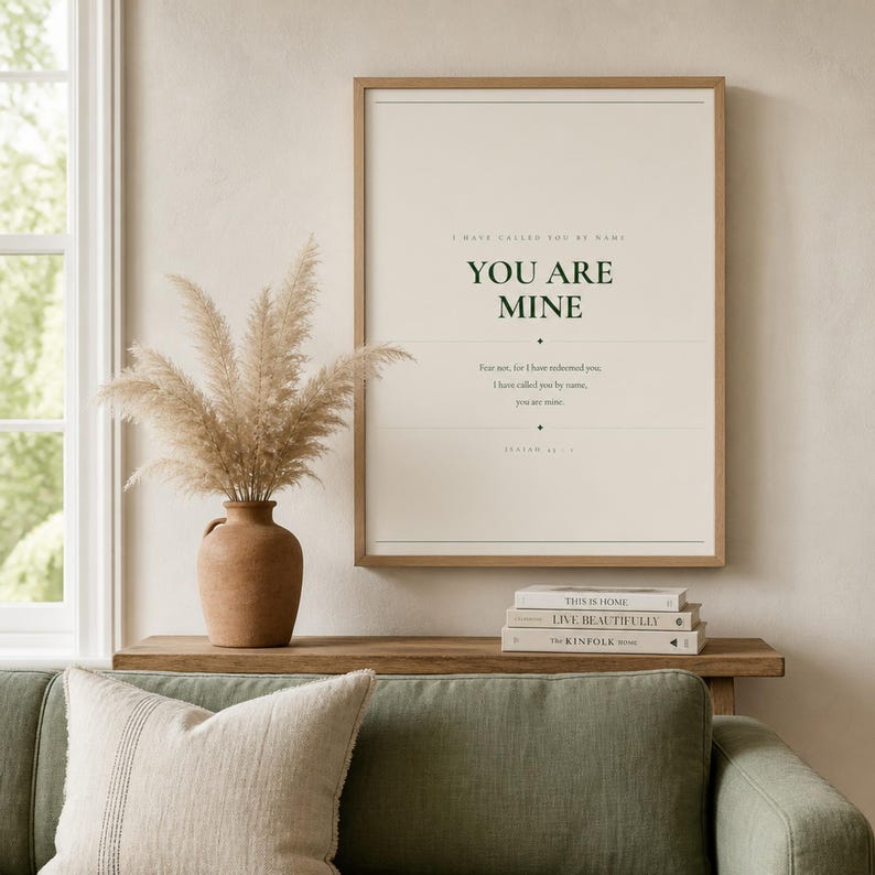 Warm lifestyle room with the framed design on a soft cream wall beside neutral decor and natural window light. The You Are Mine artwork features Isaiah 43:1 in minimalist forest green typography on warm beige. This Isaiah 43:1 print suits a nursery, baptism gift, baby dedication, bedroom, or calm Christian reading corner as printable digital wall art.