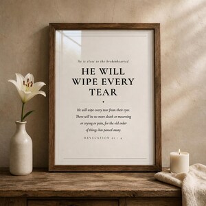 Airy room scene with the framed design above comfortable neutral furniture in a calm, modern Christian home setting. The He Will Wipe Every Tear artwork presents Revelation 21:4 in minimalist black typography on warm beige. This Revelation 21:4 print suits a sympathy gift, memorial space, prayer room, bedroom, or comforting Christian home as printable digital wall art.