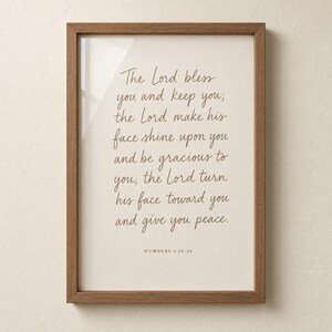 Hero close-up of the Numbers 6:24-26 Aaronic blessing print in a warm honey-oak frame with clear glass reflection on a cream plaster wall. Hand-lettered terracotta typography 'The Lord bless you and keep you' on cream paper. Premium modern Christian wedding scripture wall art for instant digital download.