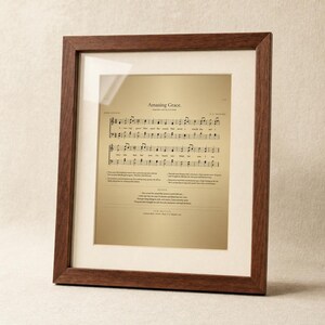 Amazing Grace antique hymnal page printable wall art in a thin walnut hardwood frame on a soft cream plaster wall — vintage sheet music notation with NEW BRITAIN tune and John Newton 1779 credit. Editorial product shot of Christian hymn print, instant digital download from TheFramedVerse on Etsy.