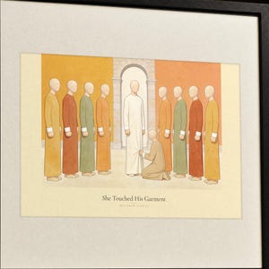 Angled close-up of the She Touched His Garment Matthew 9:20-22 print in its walnut frame, showing the cream mat edge, fine wood grain, and the soft watercolor-style illustration in warm terracotta, ochre and sage tones with hand-lettered verse type, biblical wall art detail shot, Christian home decor digital download