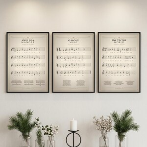Christmas Hymns Set of 3 vintage hymn prints — Away in a Manger, O Holy Night, Joy to the World — in thin black metal frames, frontal view on off-white textured wall, modern classical Christmas gallery wall arrangement, soft top light with gentle shadow, holiday christian wall art digital download TheFramedVerse