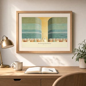 Desk mockup of The Parting of the Red Sea in a light oak frame above a wooden workspace with a brass lamp, ceramic mug, open notebook, pen, and leafy plant. The artwork shows Moses on the centre axis between two towering walls of water while the people cross the dry seabed. Angled sunlight, neutral styling, and cream matting place the Bible story print in a quiet study or home office setting.