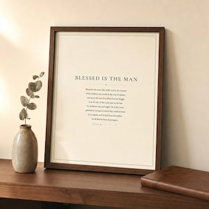 Styled shelfie: Psalm 1 Blessed Is The Man art print leaning in a walnut frame on a wooden shelf alongside a leather-bound bible, a small green plant, and a ceramic mug. Warm lifestyle scene showing the forest green and cream scripture print in a cozy home setting. Christian Father's Day gift for dad, Psalm 1:1-3 bible verse wall art.