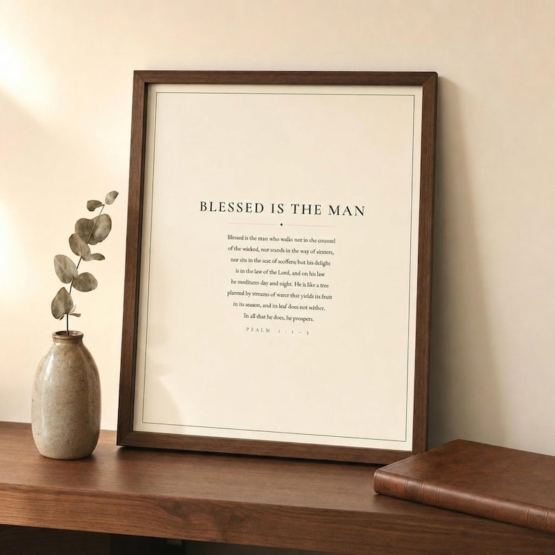 Styled shelfie: Psalm 1 Blessed Is The Man art print leaning in a walnut frame on a wooden shelf alongside a leather-bound bible, a small green plant, and a ceramic mug. Warm lifestyle scene showing the forest green and cream scripture print in a cozy home setting. Christian Father's Day gift for dad, Psalm 1:1-3 bible verse wall art.