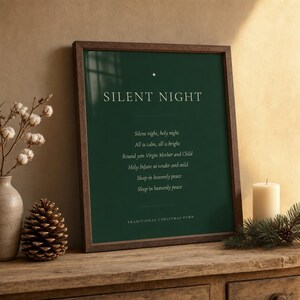 Silent Night Christmas print in a walnut frame resting on a wooden shelf beside a glowing candle and evergreen sprig. Cream serif lettering and a gold star ornament stand out on the deep forest green artwork, with the first hymn verse centered below. A cozy Christmas hymn wall art display for understated Christian Christmas decor and seasonal styling.