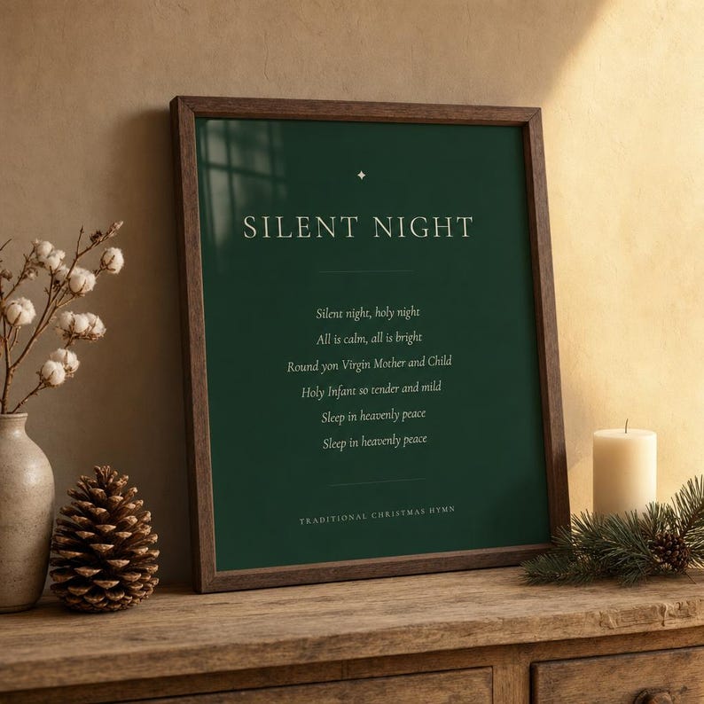 Silent Night Christmas print in a walnut frame resting on a wooden shelf beside a glowing candle and evergreen sprig. Cream serif lettering and a gold star ornament stand out on the deep forest green artwork, with the first hymn verse centered below. A cozy Christmas hymn wall art display for understated Christian Christmas decor and seasonal styling.
