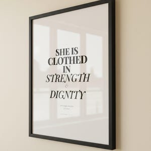 Proverbs 31 25 Wall Art She is Clothed in Strength and Dignity Scripture Print, — "She is clothed with strength and dignity, and she laughs without fear of the future", in elegant Mother's Day design with soft pink watercolor botanical flowers and graceful serif typography, presented as an elegant flat lay on white marble with a fountain pen and eucalyptus sprig