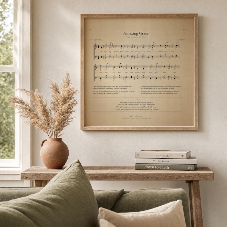 Amazing Grace vintage hymnal page printable in a slim warm-walnut frame, leaning on white Carrara marble next to a clear glass bud vase with a single fresh olive branch and an open vintage leather hymnal showing music notation pages. Premium Pinterest-worthy Christian sheet music print, instant digital download from TheFramedVerse on Etsy.
