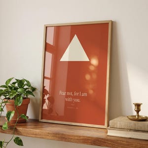 Isaiah 41:10 Fear not for I am with you trendy christian wall art in oak glass frame on wooden shelf with pothos plant, vintage books and brass candle holder. Terracotta red modern maximalist bible verse print with cream triangle shape, warm window l