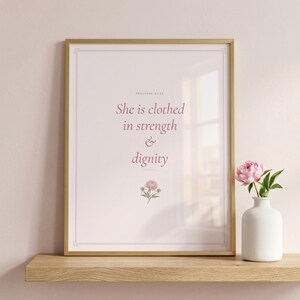 Coquette Christian wall art closeup of She Is Clothed In Strength And Dignity Proverbs 31:25 scripture print in pink blush coquette style with peony watercolor, displayed in elegant gold frame with glass reflection on light oak shelf with fresh pink peony in white vase, soft warm lighting
