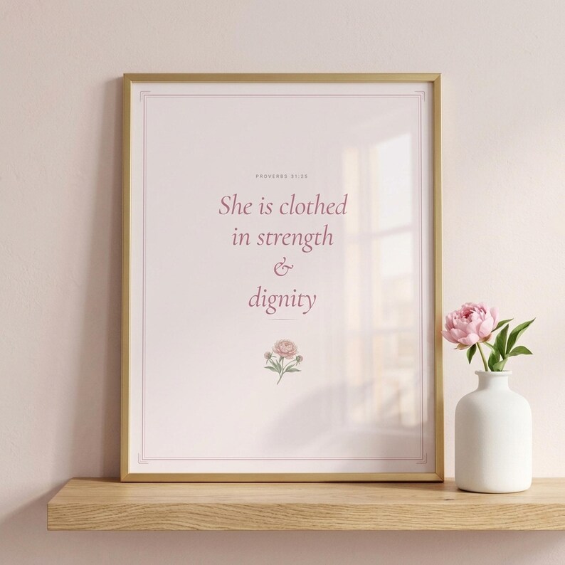 Coquette Christian wall art closeup of She Is Clothed In Strength And Dignity Proverbs 31:25 scripture print in pink blush coquette style with peony watercolor, displayed in elegant gold frame with glass reflection on light oak shelf with fresh pink peony in white vase, soft warm lighting