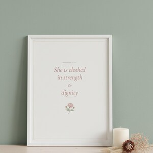She Is Clothed in Strength and Dignity Proverbs 31 25 Wall Art Coquette Christia — lifestyle mockup in modern room setting with elegant framed print on wall. This coquette bow, botanical, pink blush christian scripture wall art print features beautiful serif typography on warm cream background. Proverbs 31 25 bible verse digital download printable for home office bedroom living room gallery wal...