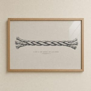 Front view of the Cord of Three Strands print in a warm oak wood frame on a cream wall, glass reflection visible. Pencil-sketch braided rope illustration with Ecclesiastes 4:12 reference. Modern Christian wedding bible verse wall art for instant digital download.