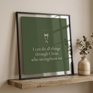 Coquette Christian wall art closeup of I Can Do All Things Through Christ Philippians 4:13 scripture print in sage green coquette style with satin ribbon bow, displayed in sleek black metal frame with glass reflection on wooden floating shelf with eucalyptus in ceramic vase, warm natural light