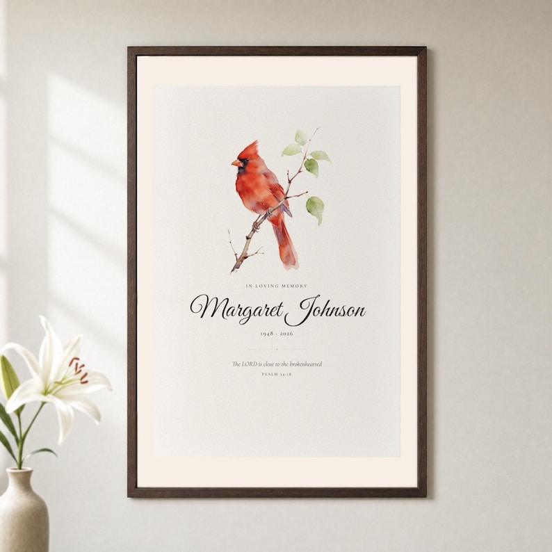 Hero framed view of a personalized Christian memorial print with a detailed red cardinal resting near delicate branches. In Loving Memory appears above the custom name Margaret Johnson and the dates 1948 to 2026, followed by Psalm 34:18 and The Lord is close to the brokenhearted. Clear glass and a natural frame reveal the gentle paper texture and readable tribute.