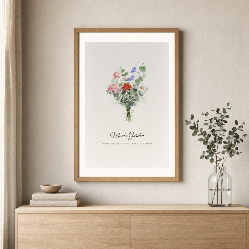 Lifestyle room mockup showing a personalized birth flower bouquet in a light wood frame above a warm neutral console. The watercolor family flower art includes individual birth month flowers, names and the custom title Mom's Garden. Soft daylight, a ceramic vase and simple decor show how this Christmas gift for mom or grandma looks in a calm modern home.