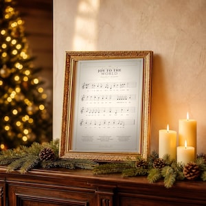 Joy to the World vintage hymn print featuring Psalm 98:4 framed in light oak resting on a wooden console with stacked books, pinecones and dried eucalyptus by a sunlit window, cozy farmhouse living room. Single Christmas sheet music wall art digital download from TheFramedVerse Etsy shop, aged sepia hymnal page style in cream and soft black tones.