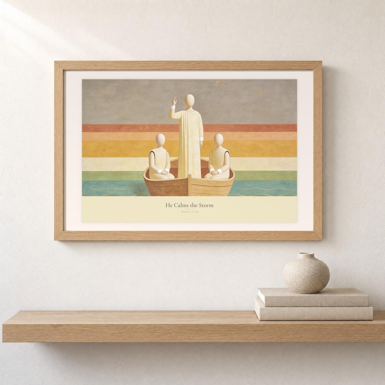 Shelf mockup of He Calms the Storm in a light oak frame resting on a pale wood surface beside stacked neutral books and a rounded ceramic vase. The framed print shows Christ standing in a wooden boat with a raised hand above calm layered water. Cream matting, soft wall texture, and natural light present the horizontal Bible artwork at an easy domestic scale.