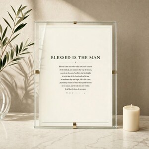 Psalm 1 Blessed Is The Man scripture art print displayed in a clear glass frame with soft light reflections on a white marble surface beside eucalyptus sprigs and a cream candle. Elegant minimal styling, dark forest green typography on cream, evoking calm and faith. Printable christian wall art, Father's Day bible verse gift for dad.