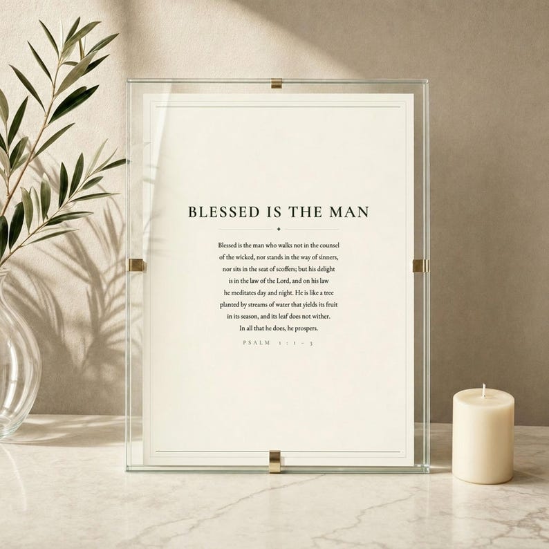 Psalm 1 Blessed Is The Man scripture art print displayed in a clear glass frame with soft light reflections on a white marble surface beside eucalyptus sprigs and a cream candle. Elegant minimal styling, dark forest green typography on cream, evoking calm and faith. Printable christian wall art, Father's Day bible verse gift for dad.
