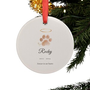Lifestyle scene showing a round personalized acrylic pet memorial ornament hanging from a ribbon on a softly lit Christmas tree. The watercolor Paw with Halo design includes the pet name Rocky and the years 2012 to 2024. Warm lights and evergreen branches frame the remembrance keepsake while the ornament remains large, clear and easy to see.