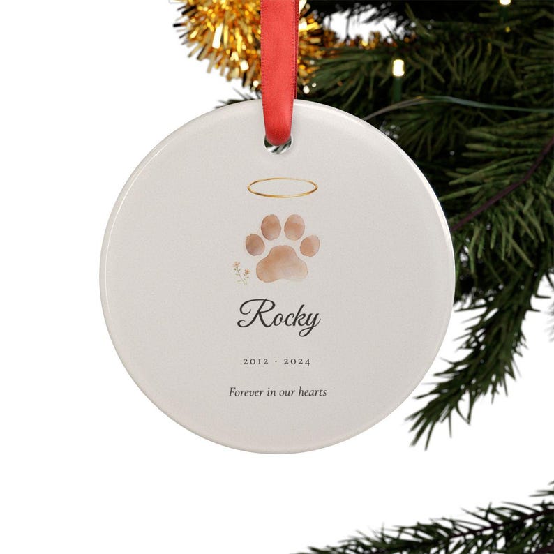 Lifestyle scene showing a round personalized acrylic pet memorial ornament hanging from a ribbon on a softly lit Christmas tree. The watercolor Paw with Halo design includes the pet name Rocky and the years 2012 to 2024. Warm lights and evergreen branches frame the remembrance keepsake while the ornament remains large, clear and easy to see.